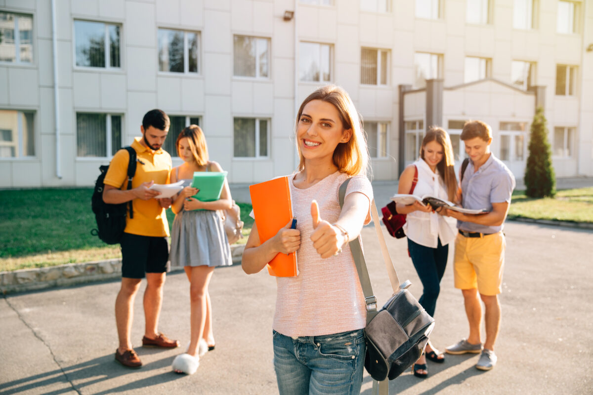Jeune femme le pouce levé devant un établissement scolaires, avec d'autres jeunes personnes derrière elle