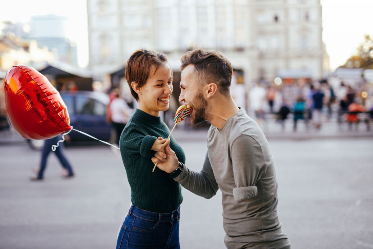 Un couple se sourit. Tous deux tiennent une sucette en forme de cœur. La femme a un bras raccourci. L'homme n'a pas de bras à gauche.