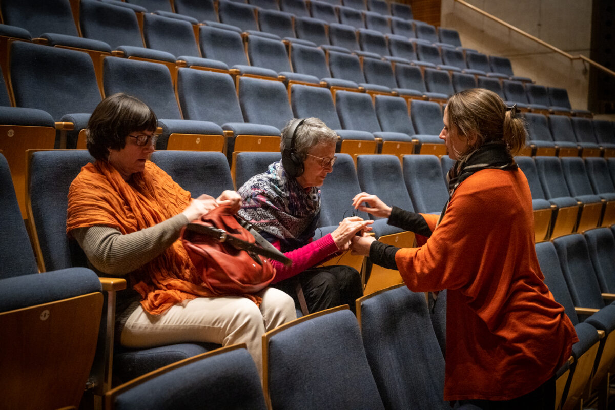 Dans les gradins d’un théâtre deux femmes sont assises, l’une d’elle porte un casque audio. Face à elles, une troisième femme, agenouillée sur un siège, explique le maniement du boîtier pour régler le volume du son.