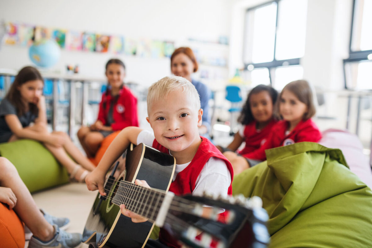 Un enfant souriant i tient une guitare à la main. L'enfant est assis dans un groupe de plusieurs enfants et d'un adulte.